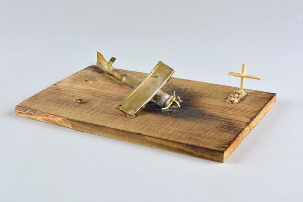 This model of a plane crash site mounted on a wooden board. There is a small crashed plane taking up most of the board with a small model of a grave to one end and side.