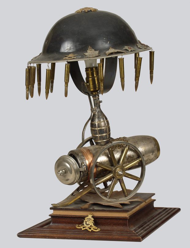 A metallic exotic looking table lamp constructed from various war implements. A war helmet surrounded by dangling bullets is the shade for a lamp body built from spent ammunition.