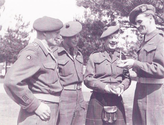A sepia photograph of three soldiers looking at a crafted ashtray held by the forth. All four men are smiling and standing outdoors.