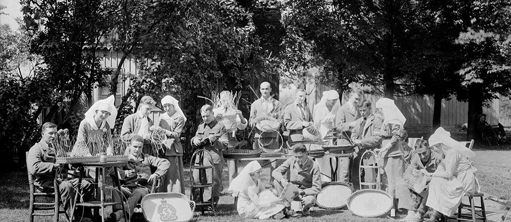 A black and white photo of several recovering First World War veterans with their nurses. They are outdoors on a bright day surrounding tables holding their crafted items.