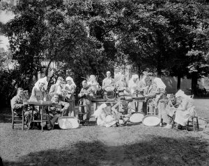 A black and white photo of several recovering First World War veterans with their nurses. They are outdoors on a bright day surrounding tables holding their crafted items.