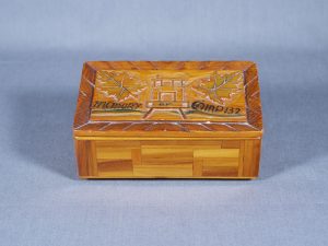 Petite boîte de bois faite à la main, avec couvercle sculpté montrant une maisonnette, avec une feuille d’érable de chaque côté.