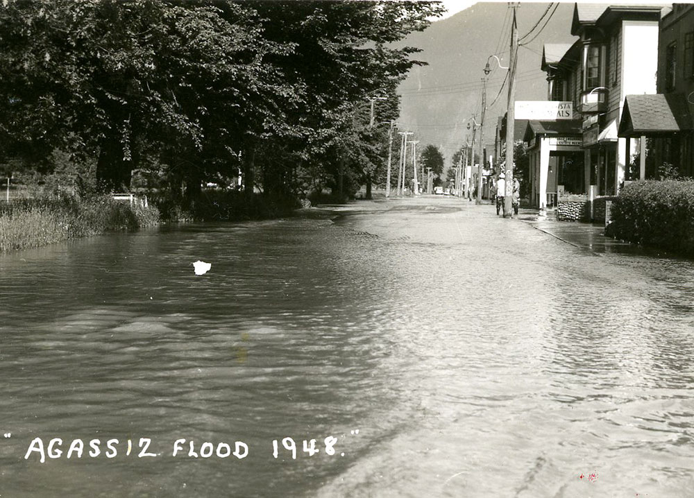 Black and white photograph of a flooded street with buildings on one side and trees on the other.
