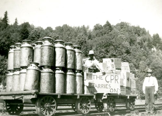 Black and white photograph with milk cans stacked on a flatbed rail cart. Two men hold a sign The CPR Carries On.
