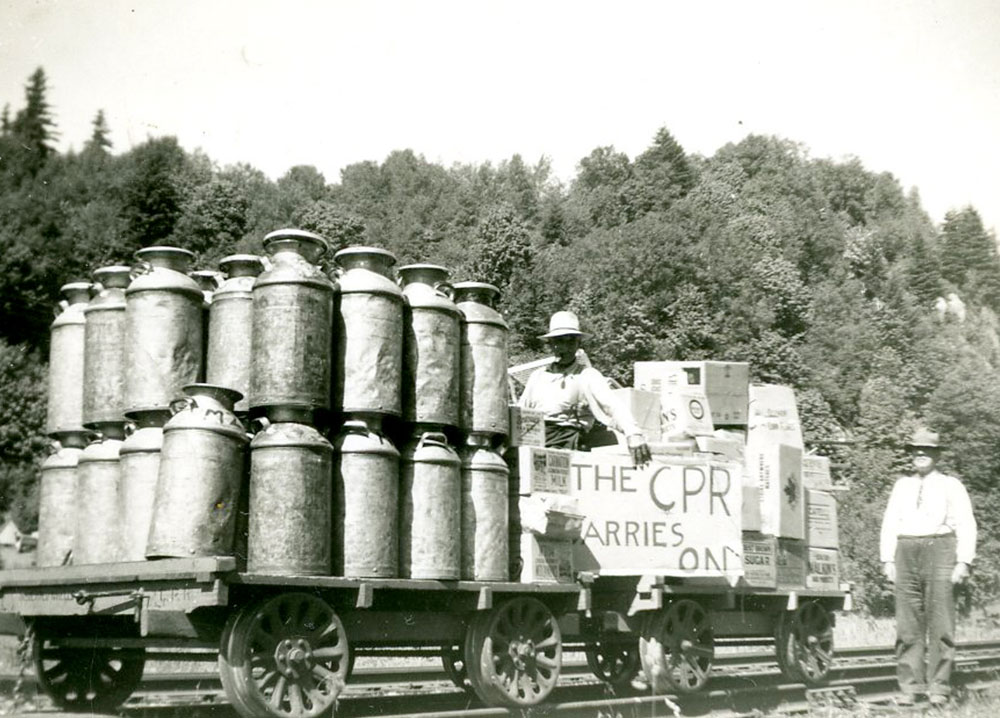 Black and white photograph with milk cans stacked on a flatbed rail cart. Two men hold a sign "The CPR Carries On".