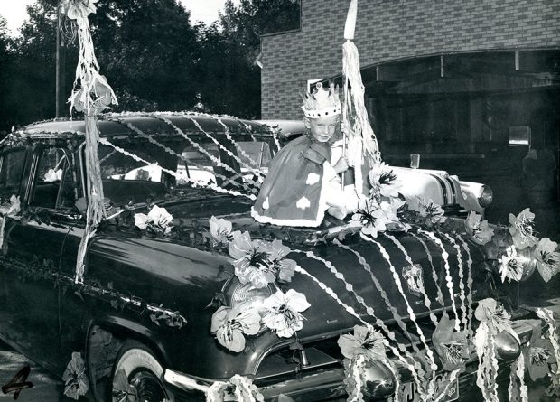 Black and white photograph of a young boy wearing a robe and a crown and sitting on the hood of a car, which is decorated for a parade.
