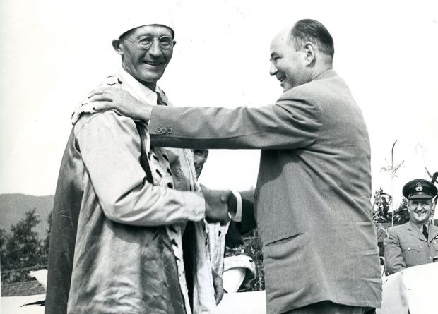 Black and white photograph of two men standing on a stage, shaking hands. One man is wearing a robe and a crown while the other wears a suit, 1954.