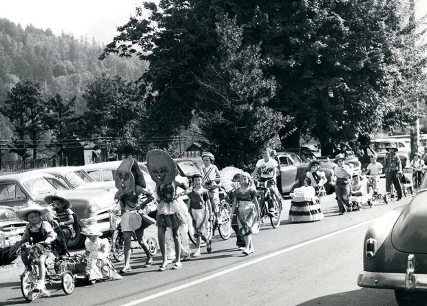 Black and white photograph of children in costumes, walking and riding bikes on a street as part of a parade, 1953.