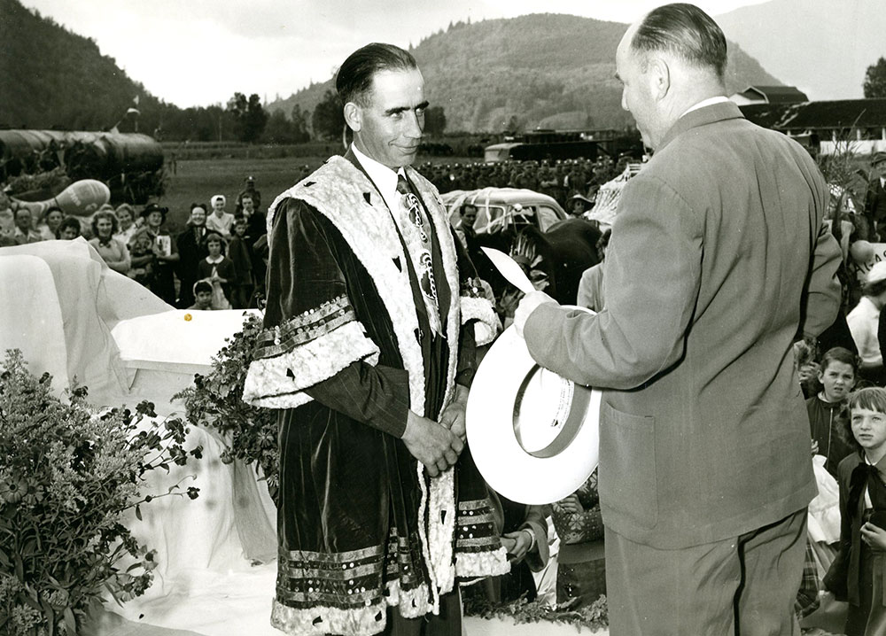 Black and white photograph of two men standing on a stage. One is wearing a robe and the other is wearing a suit and holding a cheque, 1953.