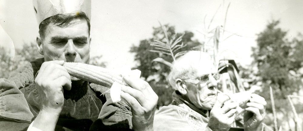 Black and white photograph of two men dressed in robes eating corn on the cob. The man on the left is 1950 Corn King Norman Morrow. The man on the right is 1949 Corn King Sam Stock.