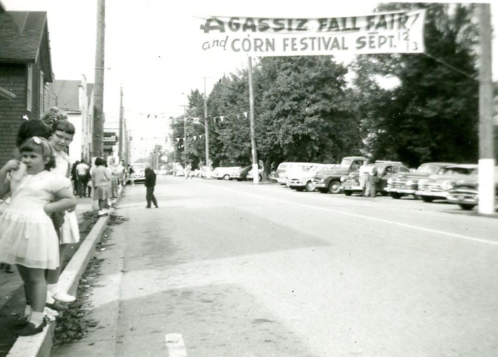 Black and white photograph of people gathered along a street to watch a parade. A banner hangs over the street "Agassiz Fall Fair and Corn Festival, Sept. 12 and 13."