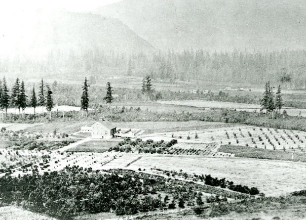 Black and white photograph of the Agassiz Dominion Experimental Farm. It is surrounded by fields, trees, and mountains.
