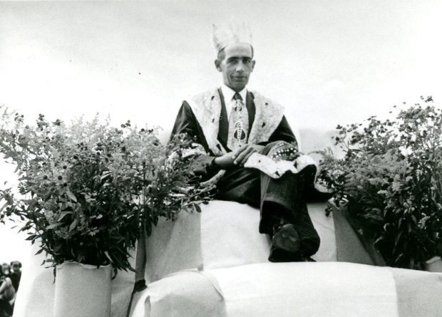 Black and white photograph of a man wearing a robe and a crown and sitting on a throne surrounded by flowers. He is the 1951 Corn King.