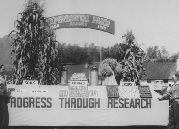 Black and white photograph of a farm display. There is an arch in the background with two high stalks of corn on each side naming the Experimental Farm Agassiz, 1888 to 1959. In the foreground, a table features a display of soils, animals, and crops with a sign hung on the front of the table promoting Progress through Research.