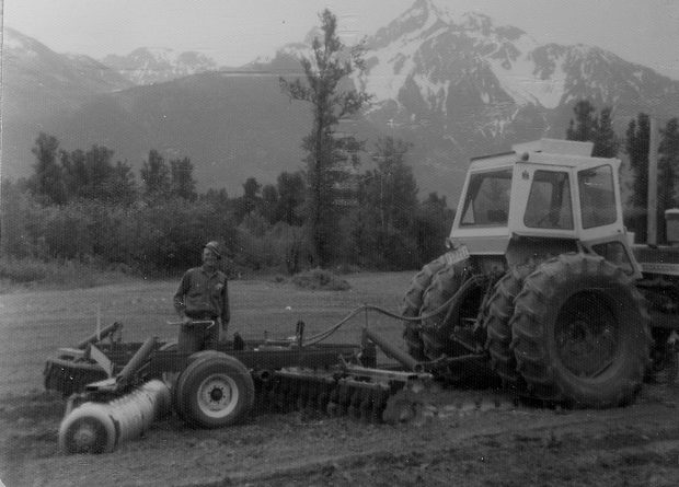 Black and white photograph of a man with a tractor pulling discing equipment in a field and Mount Cheam in the background.