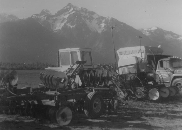 Black and white photograph of two tractors pulling discing equipment and a truck in a field. Mount Cheam is in the background.