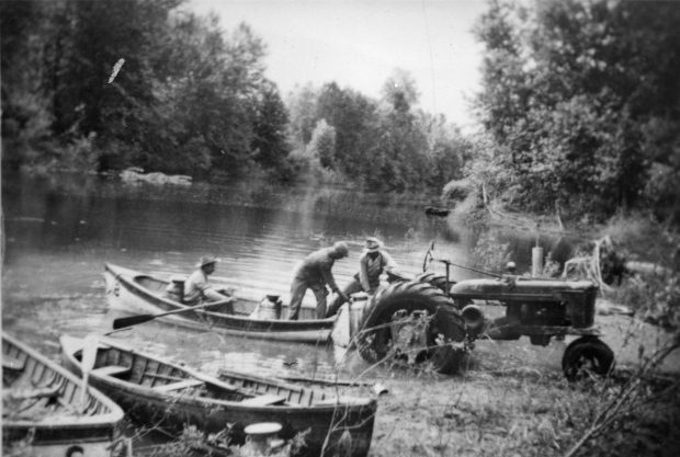 Black and white photograph of three men loading milk cannisters into a row boat from a tractor.