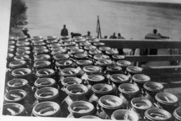 Black and white photograph of milk cannisters lined up in a truck bed at a ferry landing.
