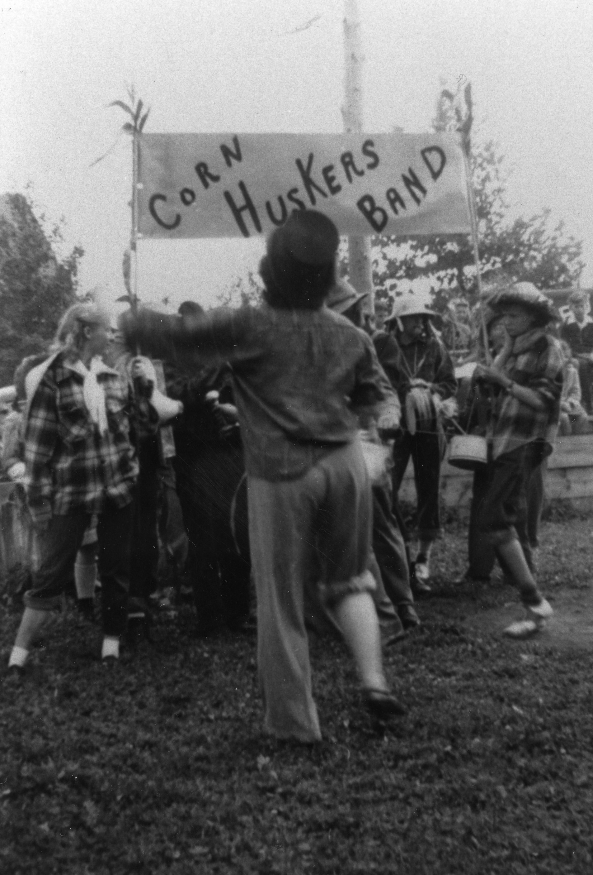 Black and white photograph of a group wearing plaid shirts and straw hats and carrying instruments. They are holding a sign "Corn Huskers Band."
