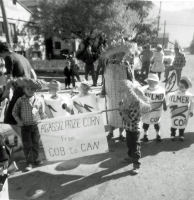 Black and white photograph of children in a parade dressed in corn cob and corn can costumes. Two children are holding a sign Agassiz Prize Corn from Cob to Can.