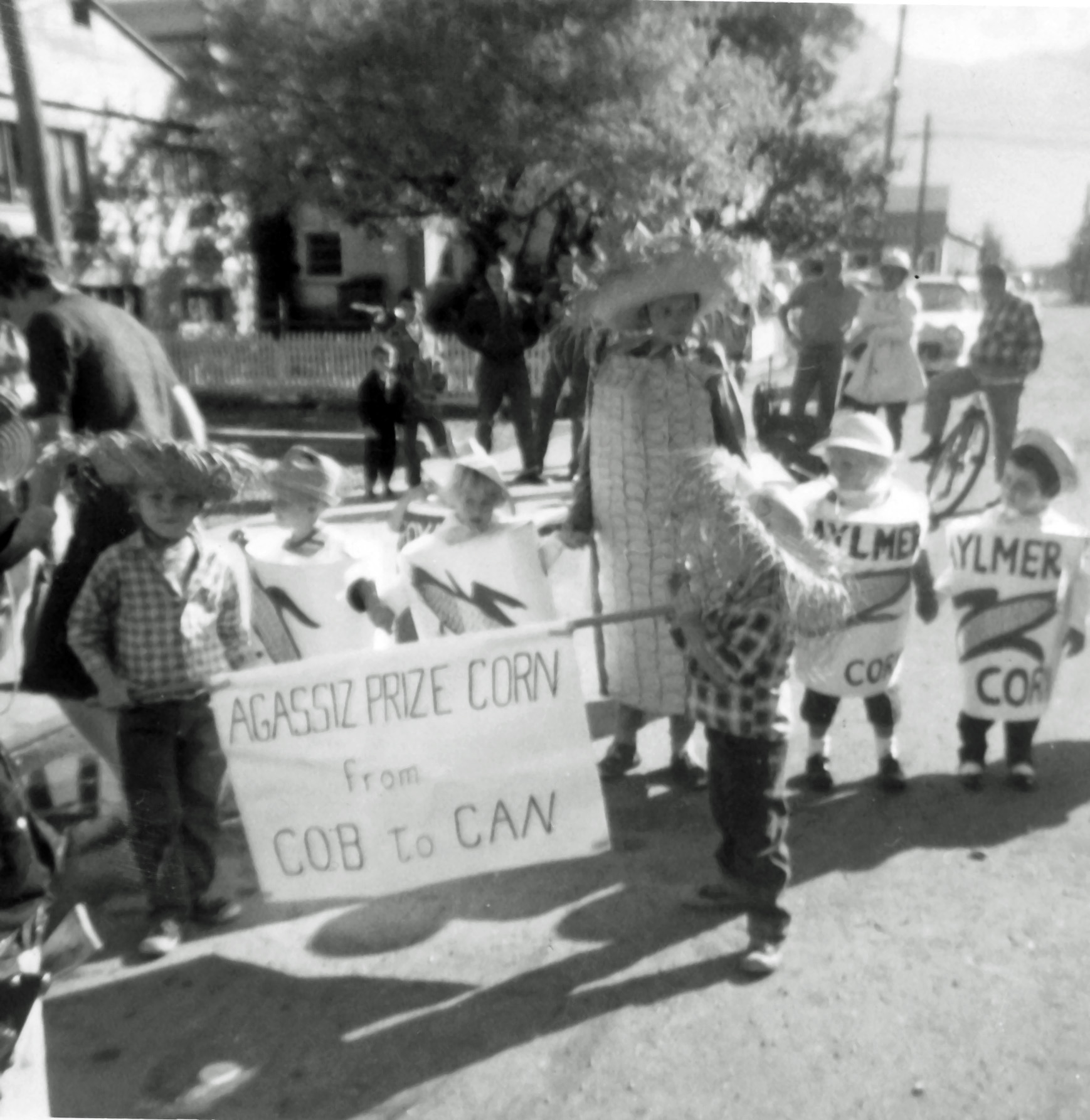 Black and white photograph of children in a parade dressed in corn cob and corn can costumes. Two children are holding a sign "Agassiz Prize Corn from Cob to Can."