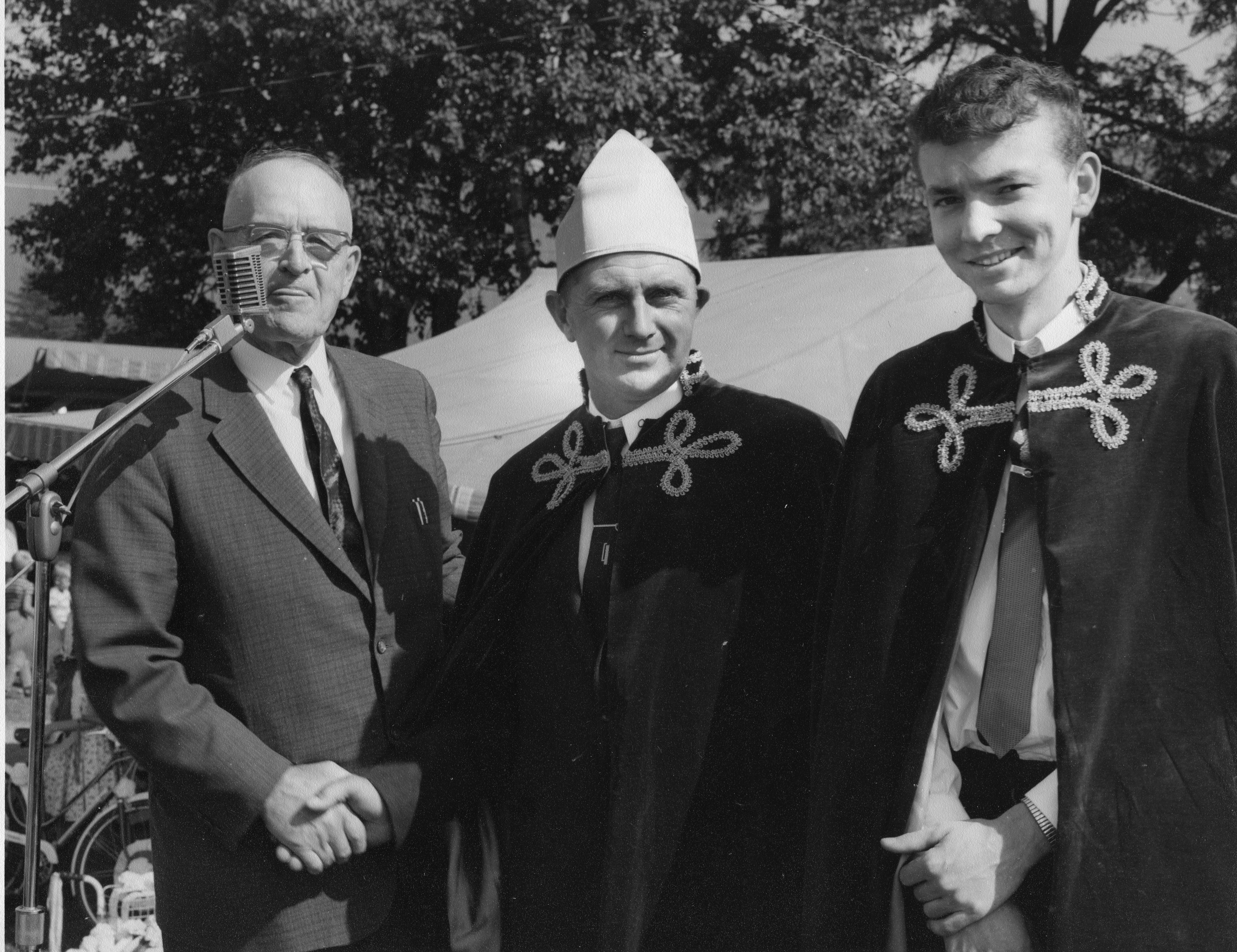 Black and white photograph of three men: one man is wearing a suit and the other two are wearing robes. The two men on the right are the corn kings from 1963 and 1964. The man on the left in front of the microphone is the master of ceremonies.