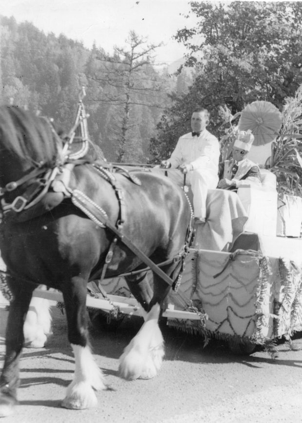 Black and white photograph of a horse team pulling a float in a parade. The driver is dressed in a white suit and the passenger is wearing a robe and a crown. He is the 1951 Corn King.