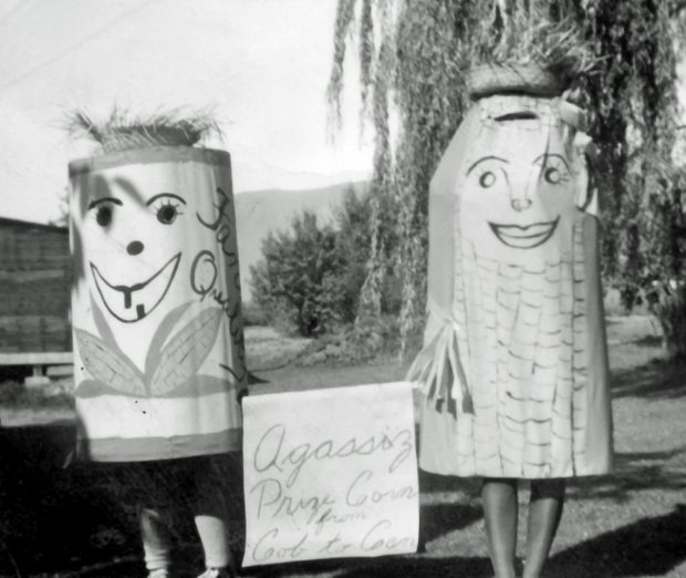 Black and white photograph of two people in corn cob and can of corn costumes. They are holding a sign Agassiz Prize Corn from Cob to Can.