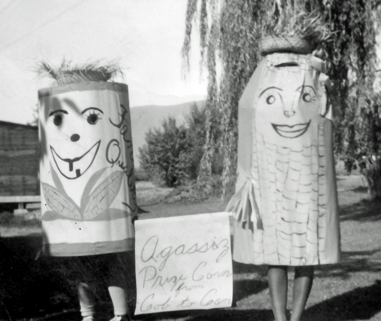 Black and white photograph of two people in corn cob and can of corn costumes. They are holding a sign "Agassiz Prize Corn from Cob to Can."