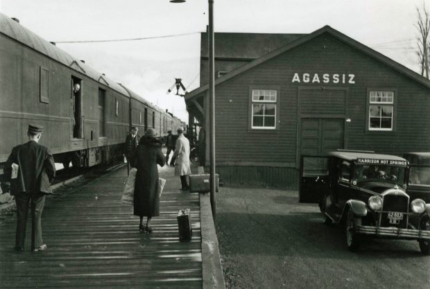 Black and white photograph of a train at the Agassiz station with passengers waiting on a platform. A Harrison Hot Springs taxi is in the foreground.
