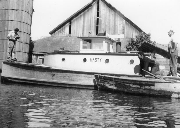 Black and white photograph of three men loading milk onto the Hasty tugboat in front of a barn.