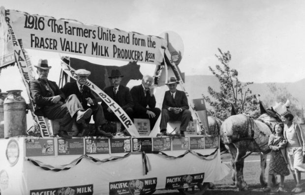 Black and white photograph of five men on a parade float pulled by a team of horses. There is a sign on the float, 1916 The Farmers Unite and form the Fraser Valley Milk Producers Assn. They are promoting Fraser Valley cheese, butter, and milk.