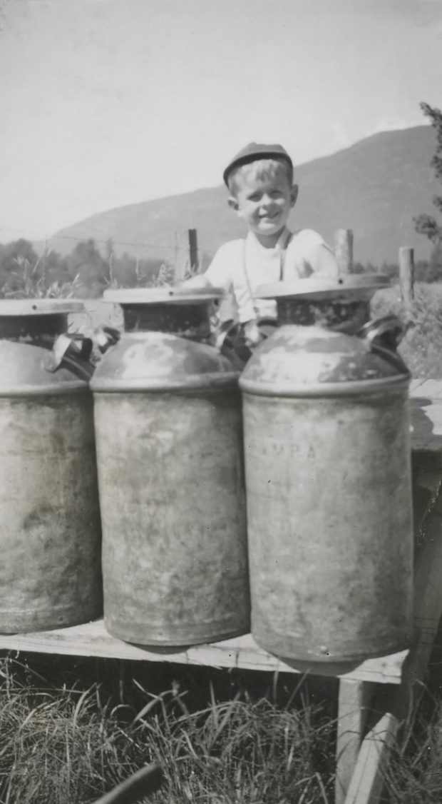 Black and white photograph of a boy with cannisters of milk on a wood stand.
