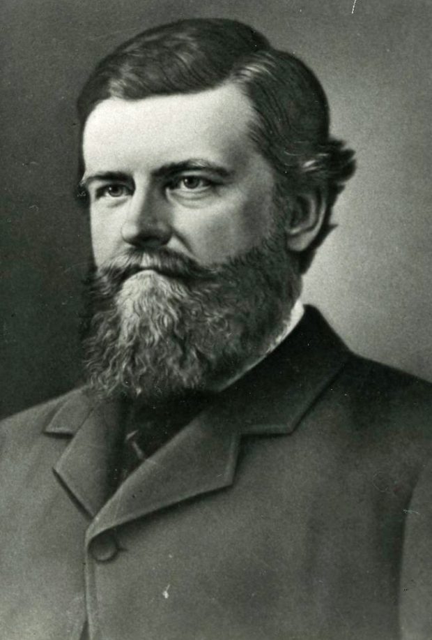 Black and white photograph portrait of a dark haired man with a beard and moustache. He is wearing a dress coat and tie.
