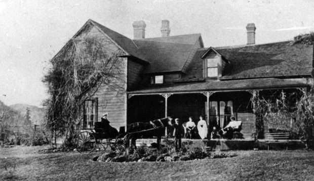 Black and white photograph of a farm house. Women are sitting on the porch looking at two people in a horse-drawn carriage.