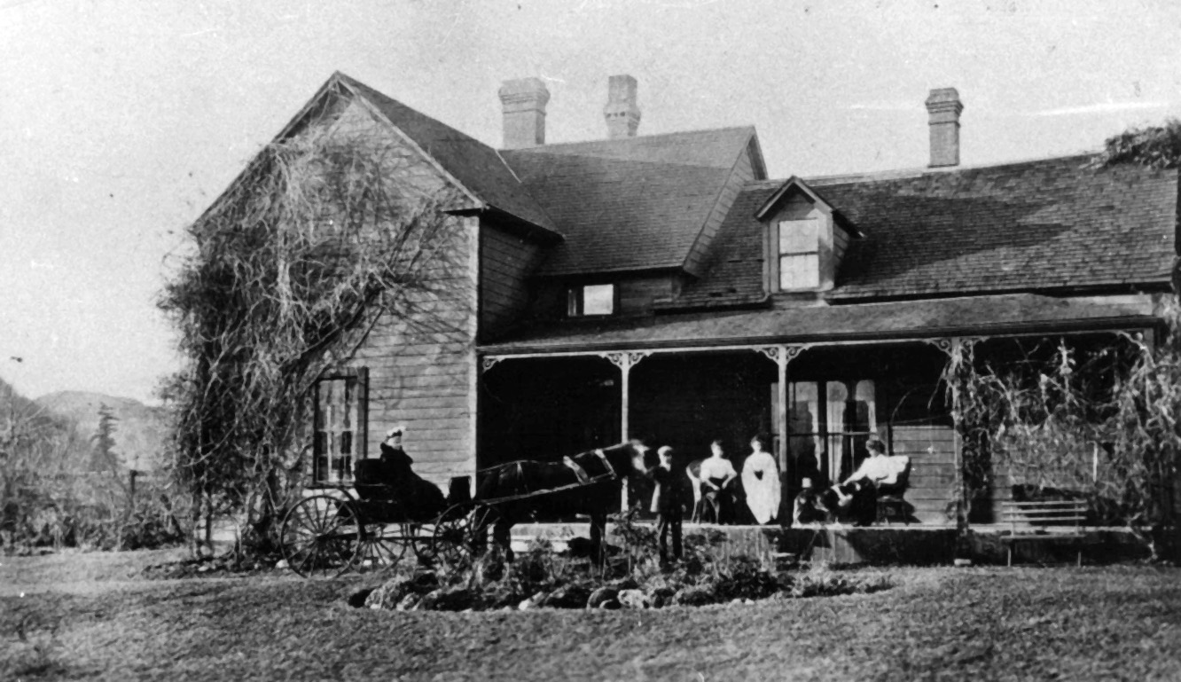 Black and white photograph of a farm house. Women are sitting on the porch looking at two people in a horse-drawn carriage.