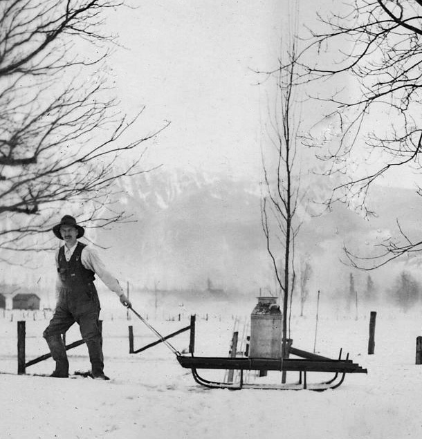 Black and white photograph of a man pulling a milk cannister on a sleigh in winter.