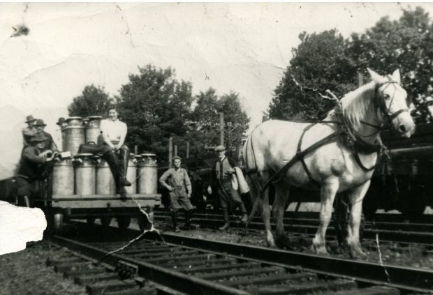 Black and white photograph of farmers with a loaded milk cart on train tracks. A work horse is attached to the cart.