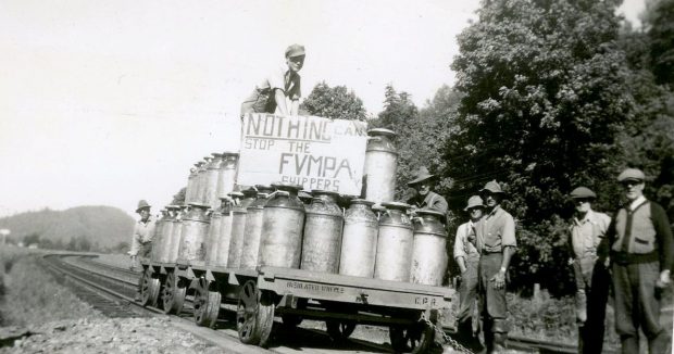 Black and white photograph of farmers with a rail cart loaded with milk cannisters. One man holds a sign Nothing can stop the FVMPA shippers.
