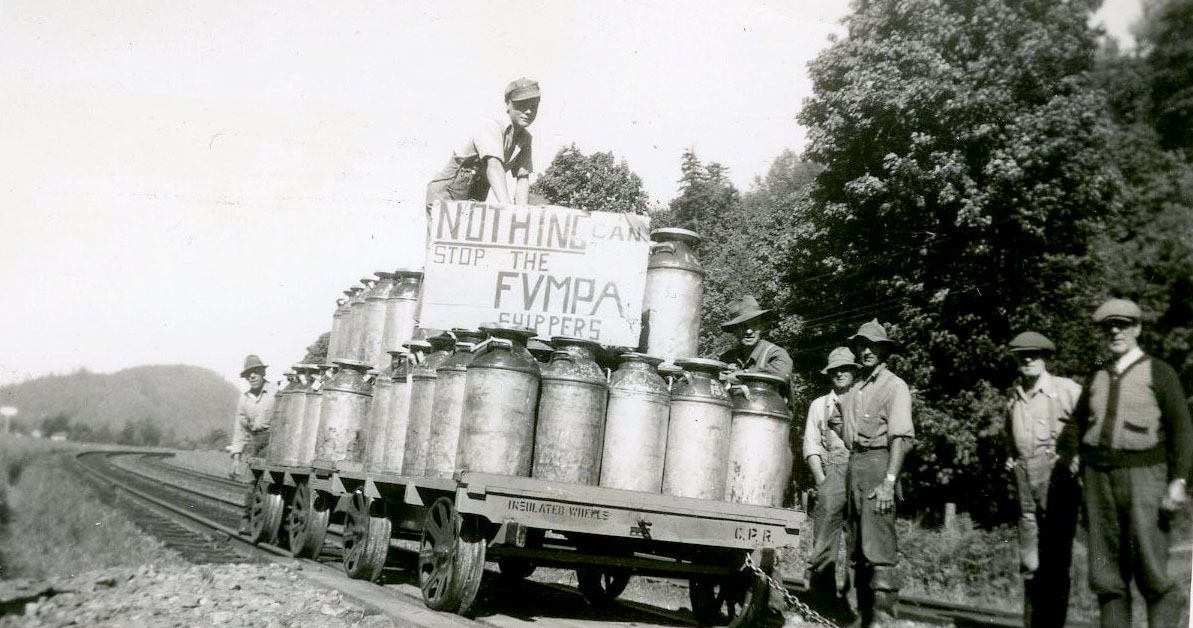 Black and white photograph of farmers with a rail cart loaded with milk cannisters. One man holds a sign "Nothing can stop the FVMPA shippers."