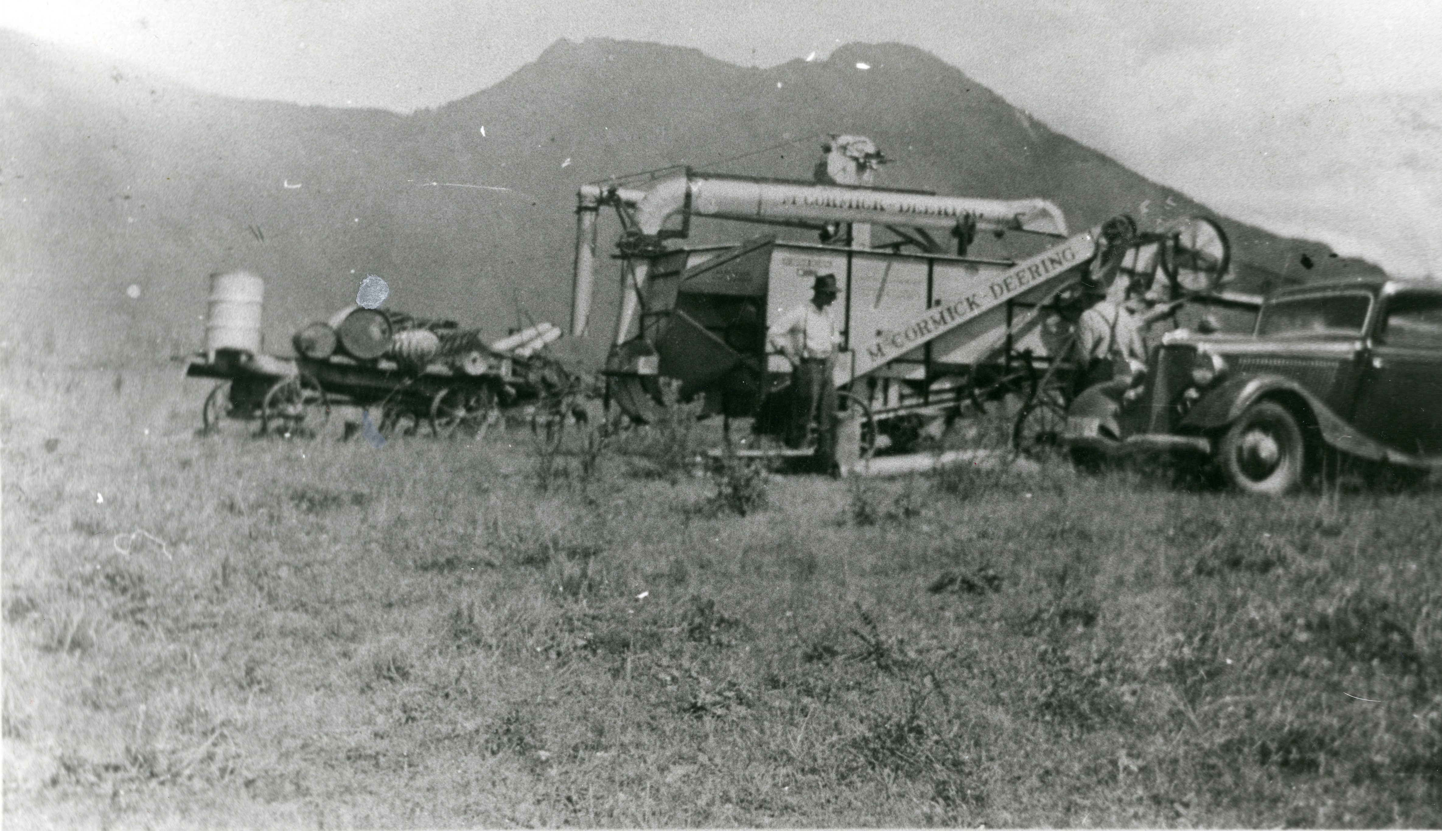 Black and white photograph of men working in a field using a McCormick-Deering thresher. A car is in the foreground and mountains are in the background. A wagon with barrels is adjacent to the thresher.