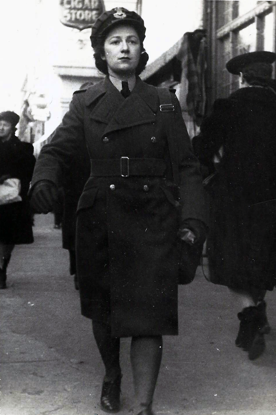 Woman in military uniform walking on busy sidewalk.