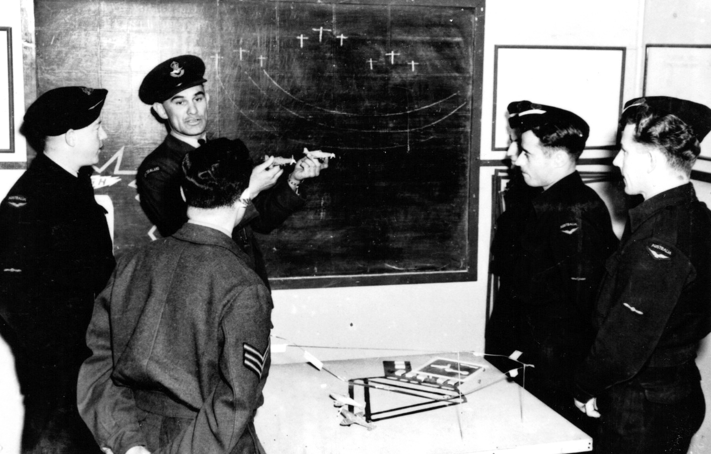 Male instructor holding two model airplanes in front of 5 students in airforce uniforms