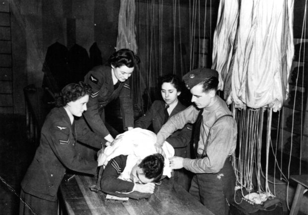 3 airwomen and one airman assisting another airman wrapped in a parachute on a table
