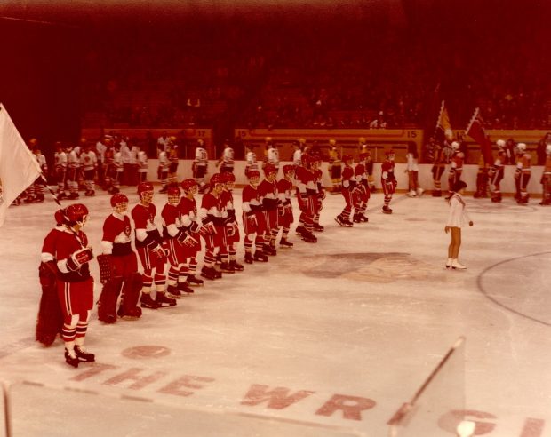 Colour photo showing a skating rink with dozens of hockey players in their uniforms. A young girl is skating in front of the boys. Spectators can be seen in the stands in the background.