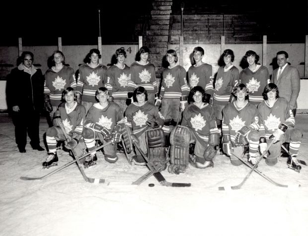 Black-and-white photo showing 14 boys in hockey uniforms, half of whom are kneeling on an ice rink with the other half standing behind them. A man is standing on each side of the boys.