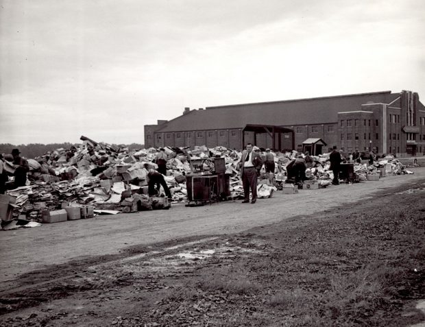 Black-and-white photo showing a collection spot located next to a building in the distance. About ten men are seen searching through this depot.