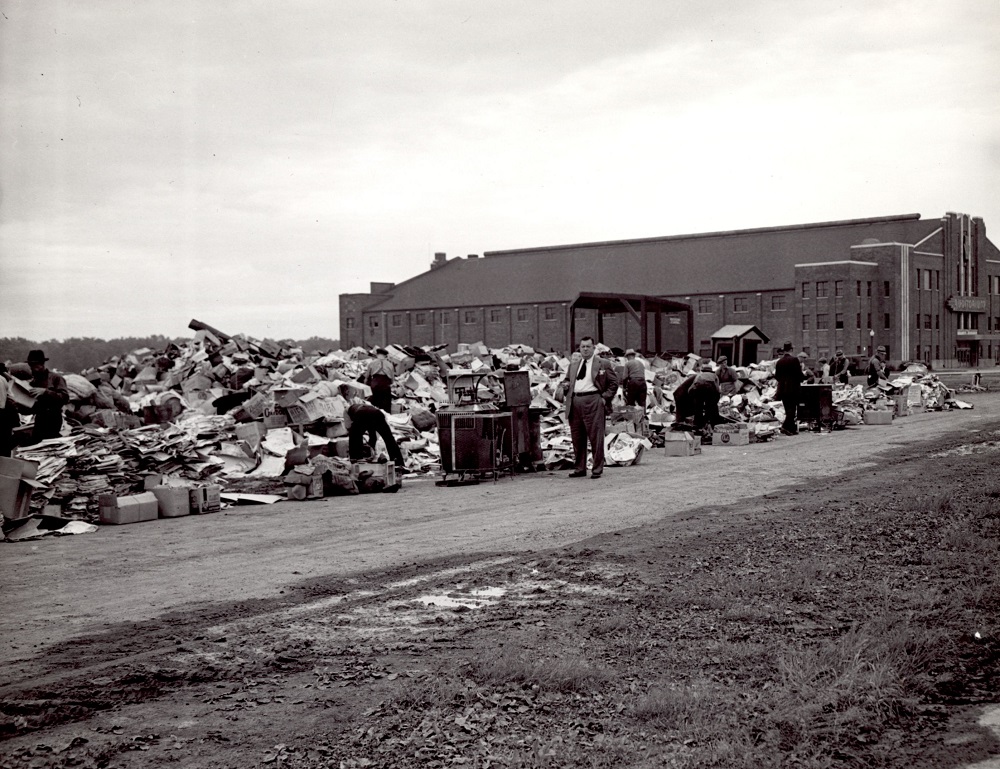 Black-and-white photo showing a collection spot located next to a building in the distance. About ten men are seen searching through this depot.