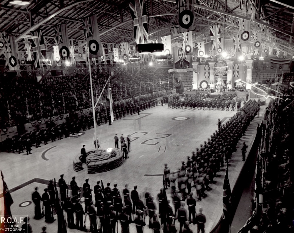 Photographie en noir et blanc où l’on voit un rassemblement de militaires à l’intérieur d’un bâtiment. Plusieurs drapeaux du Royaume-Uni flottent au-dessus de la patinoire et une foule occupe les gradins.