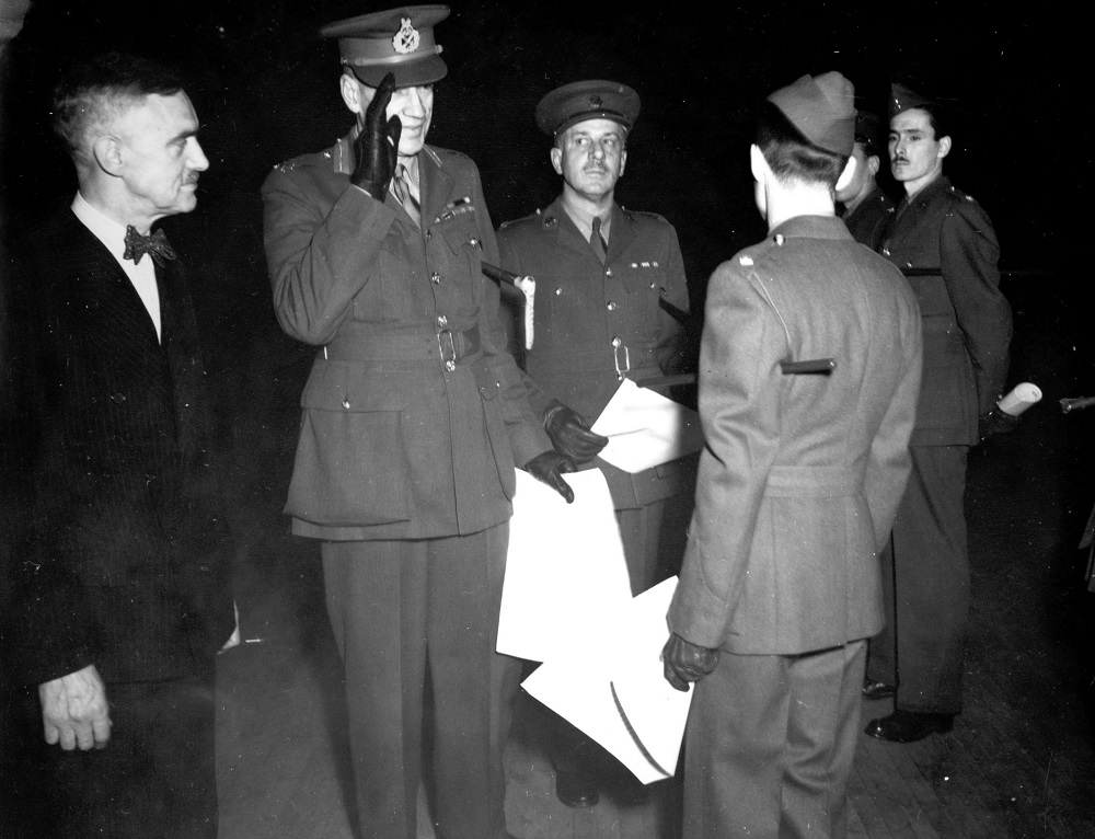 Black-and-white photo showing four servicemen in uniform and one civilian. Three soldiers are holding documents and one is giving a military salute.